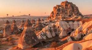 AI_IMAGE: Cappadocia fairy chimneys at sunrise with hot air balloons in the distance, ancient cave dwellings carved into volcanic rock, warm pink and gold morning light, layers of geological formations, mystical and timeless atmosphere | photorealistic | landscape
